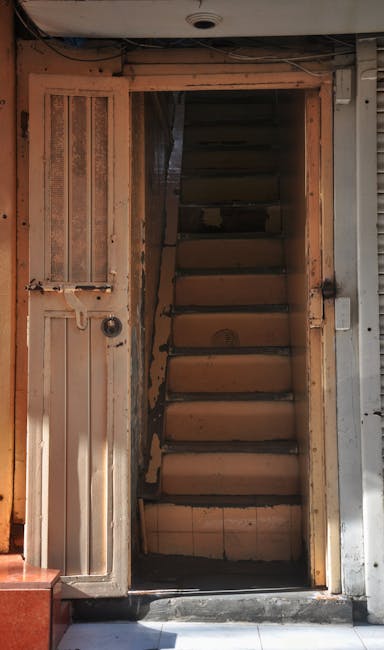 A narrow wooden staircase inside a property, with visible unfinished wooden steps and side panels, leading upward from a doorway. The doorway frame is partially constructed, revealing exposed brick and construction materials around the edges. To the left of the staircase, there is a small, closed wooden door with a window inset, also made of unfinished wood. The lighting is natural, coming from outside, casting shadows on the wooden surfaces. In the foreground, part of the ground and a small section of flooring are visible, suggesting the area is under renovation or in the process of being prepared for a move. This scene illustrates challenges in home relocation within constrained or partially renovated spaces, as addressed by Man With a Van Wapping’s removals service.
