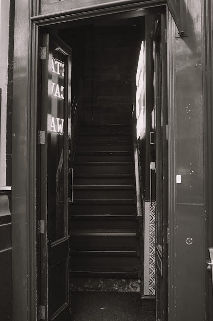 A black and white photograph showing a narrow staircase inside a building, viewed through an open doorway. The staircase has dark wooden steps with no visible handrails, and is positioned in a confined space with limited room for maneuvering large furniture. To the left side of the doorway, there is a partially visible door with a glass panel that has reflective writing on it. The surrounding wall is dark, and the lighting inside appears dim, emphasizing the tight access point. This image relates to a house relocation scenario where moving services such as Man With a Van Wapping are preparing to navigate the staircase for furniture transport. The compactness of the staircase highlights the challenge of moving in properties with restricted entry points, often requiring careful handling and strategic packing of belongings.