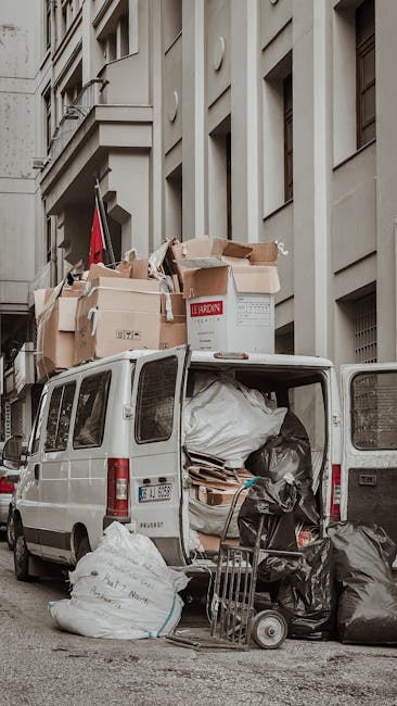 A white commercial van parked on a city street adjacent to a multi-storey residential building with a light-colored facade. The van's rear doors are open, revealing a variety of items loaded inside, including large black and white plastic garbage bags, cardboard boxes, and packaging materials. Several boxes, some labelled with red and black text, are stacked on top of the van, indicating a home relocation or furniture transport process. An industrial trolley with metal wheels is positioned next to the van, loaded with additional black garbage bags and cardboard packaging. The scene takes place during daytime with natural lighting, and the van is part of a professional removals service, such as Man With a Van Wapping, supporting furniture removals or packing and moving logistics in the Wapping area.