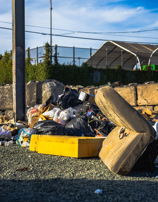 A narrow alleyway used for house removals and waste clearing, featuring a large wheeled trolley loaded with grey cardboard boxes and plastic-wrapped items positioned in the centre of the scene. Surrounding the trolley are various discarded materials, including black bin bags, scattered debris, and additional cardboard boxes. The alley is enclosed by tall, graffiti-covered brick and metal-sided walls on either side, with some graffiti and peeling paint visible. In the background, there is a pile of waste, including more cardboard and plastic wrapped items, with a tree with bare branches extending overhead. Natural light illuminates the scene, highlighting the gritty urban environment. Exterior elements such as a staircase, electrical wires, and a door frame mark entry points to nearby buildings. This image visually captures the process of loading items for a house relocation or waste disposal service, as offered by Man With a Van Wapping, in a typical urban setting suited for furniture transport, packing, and moving logistics.