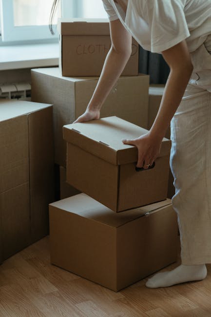 A person wearing light-colored clothing and white socks is seen stacking and lifting cardboard boxes in a well-lit room with natural light coming through a window. Several sealed cardboard boxes of varying sizes are placed on a wooden floor, with one box labeled ‘CLOTHES’ visible on top. The individual is carefully handling a medium-sized box, prepared for transportation as part of a home relocation process. The scene depicts a room inside a Wapping flat, where packing and moving preparations are underway, likely coordinated by Man With a Van Wapping. In the background, there are additional packages and furniture, indicating an organized packing and moving operation focused on furniture transport and loading logistics, with the environment organized for a professional house removal service.