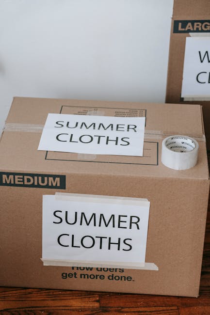 A close-up view of a cardboard moving box labeled 'SUMMER CLOTHS' in bold black letters on white tape, situated on a wooden floor inside a property. The box has a second label on the side with the same text and is sealed with clear packing tape. A roll of white adhesive tape and a smaller box marked 'LARGE' are placed on top of or behind the main box. The setting appears to be part of a home removal or packing process, with natural lighting highlighting the packing materials and furniture. The image captures a moment in the loading or packing stage of a house relocation managed by Man With a Van Wapping, focusing on organized packing for furniture transport, aligned with their removals service category.