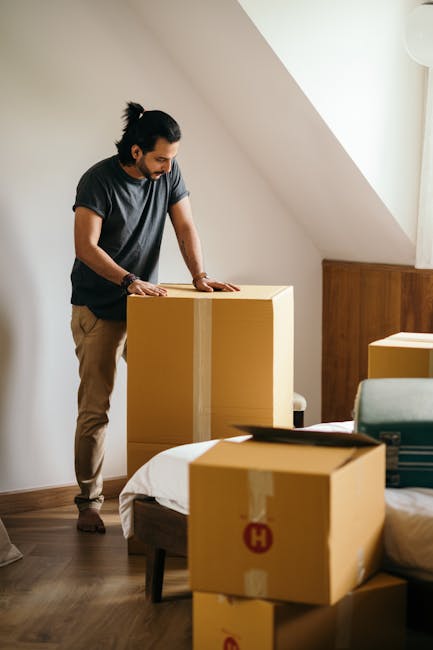 A person wearing light-colored clothing and white socks is seen stacking and lifting cardboard boxes in a well-lit room with natural light coming through a window. Several sealed cardboard boxes of varying sizes are placed on a wooden floor, with one box labeled ‘CLOTHES’ visible on top. The individual is carefully handling a medium-sized box, prepared for transportation as part of a home relocation process. The scene depicts a room inside a Wapping flat, where packing and moving preparations are underway, likely coordinated by Man With a Van Wapping. In the background, there are additional packages and furniture, indicating an organized packing and moving operation focused on furniture transport and loading logistics, with the environment organized for a professional house removal service.