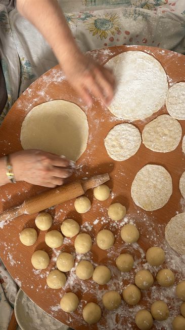 Close-up of a person's hands preparing dough on a wooden surface for home relocation packing, with circular cutouts of dough ready for baking or wrapping. The workspace is dusted with flour, and several rolled dough circles are laid out beside a rolling pin. The background shows a patterned tablecloth and some additional baking materials. The scene emphasizes careful handling and preparation of packaging materials, which can be relevant to professional removals and packing services provided by Man With a Van Wapping, supporting efficient furniture transport and moving logistics in the context of house removals.