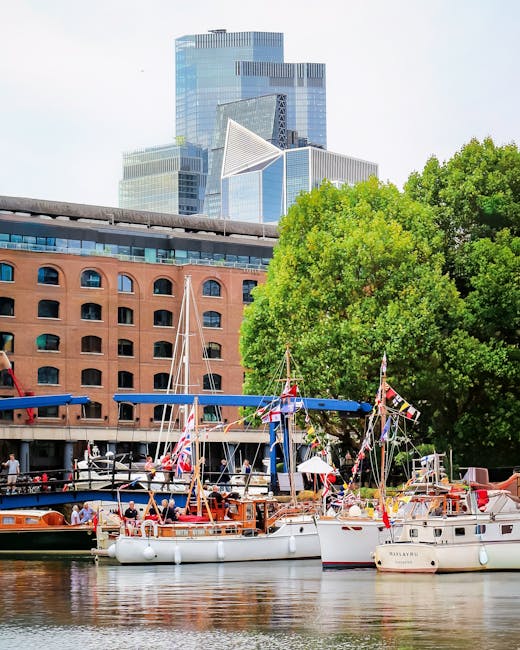 The image depicts a riverside scene with several small, wooden leisure boats moored along the quay, their decks decorated with Union Jack bunting and small flags. The boats are equipped with masts, sails, and safety equipment, and are positioned close to a retaining brick wall with a concrete walkway above. Behind the boats, there is a lush green tree providing shade, and in the background, a modern cityscape with tall glass and steel office buildings and a distinctive geometric skyscraper. The scene appears to be during daytime with bright, natural lighting, and the water reflects the boats and adjacent structures. This setting suggests a location suitable for house removals or furniture transport services, like those offered by Man With a Van Wapping, especially in relation to moving near St Katharine Docks with access to waterways and riverside environments.