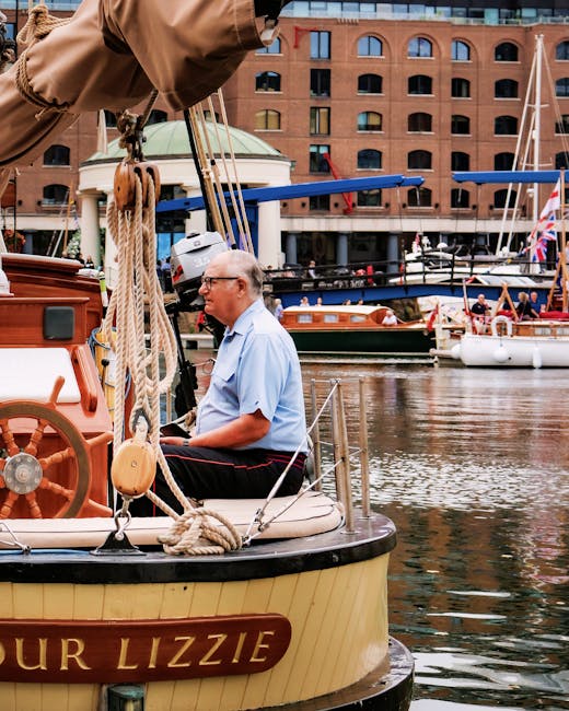 In the image, an elderly man with glasses and short white hair is seated on the deck of a small boat named 'Our Lizzie,' situated in a marina with calm water. The boat features a wooden steering wheel and is surrounded by thick, coiled ropes hanging from the mast, which is partly visible at the top of the frame. Behind him, several larger boats and yachts are moored along the waterfront, with one having a blue cover over its cockpit. In the background, there is a red brick building with multiple arched windows and balconies, indicating an urban setting near the water. The scene is lit with natural daylight, highlighting details of the boat's beige hull, the man's light blue shirt promoting a professional environment linked to house removals or moving services, and the busy, organized harbour setting. This image captures the process of boat transportation or packing at a marina, relevant to home relocation and moving logistics as managed by companies such as Man With a Van Wapping.