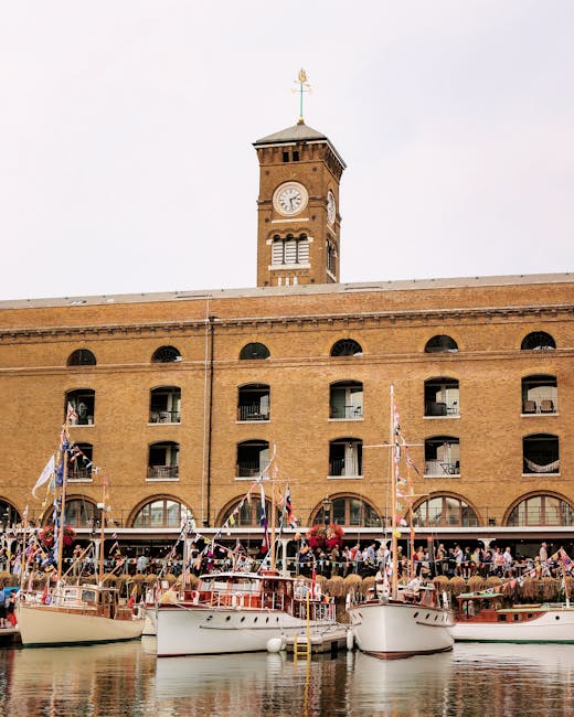 The image depicts a riverside scene with several small, wooden leisure boats moored along the quay, their decks decorated with Union Jack bunting and small flags. The boats are equipped with masts, sails, and safety equipment, and are positioned close to a retaining brick wall with a concrete walkway above. Behind the boats, there is a lush green tree providing shade, and in the background, a modern cityscape with tall glass and steel office buildings and a distinctive geometric skyscraper. The scene appears to be during daytime with bright, natural lighting, and the water reflects the boats and adjacent structures. This setting suggests a location suitable for house removals or furniture transport services, like those offered by Man With a Van Wapping, especially in relation to moving near St Katharine Docks with access to waterways and riverside environments.