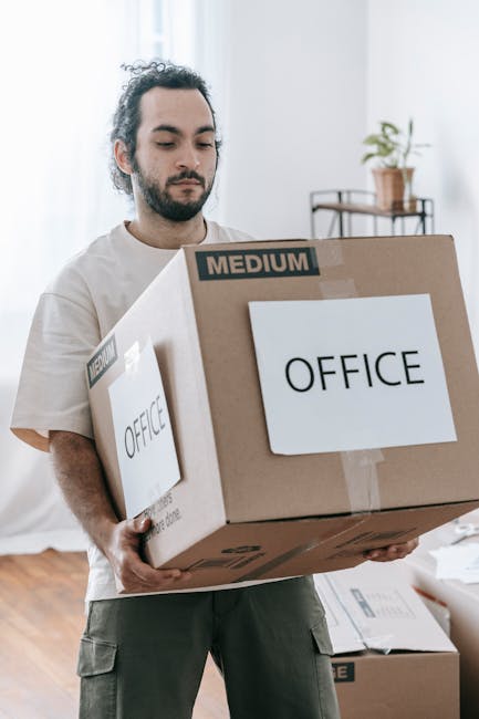 A man with dark, curly hair and a beard is inside a well-lit room, carrying a medium-sized cardboard box labeled 'OFFICE' in black text. The box appears to contain office items for a home relocation o