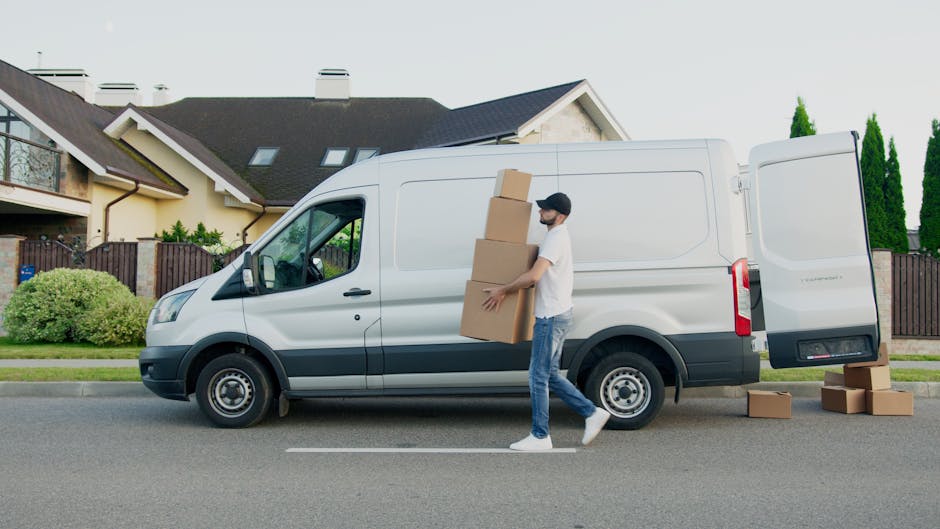 A man with a beard, wearing a black cap, white t-shirt, and blue jeans, is loading three medium-sized cardboard boxes into the side door of a white panel van parked on a residential street in Wapping,