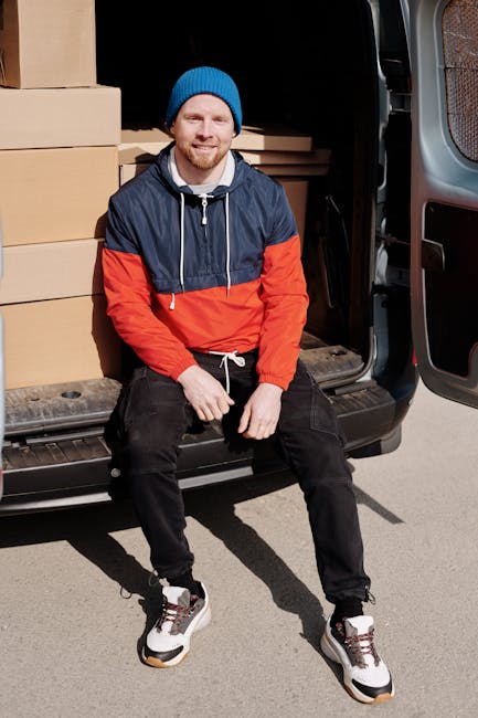 A man with a beard, wearing a black cap, white t-shirt, and blue jeans, is loading three medium-sized cardboard boxes into the side door of a white panel van parked on a residential street in Wapping,