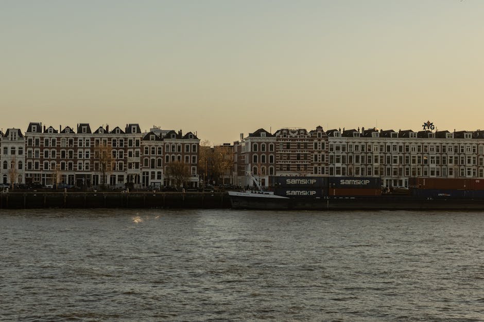 A view of a waterside residential area during dusk, featuring multiple multi-storey white and beige Victorian-style houses with black roofs, situated along a canal or riverbank. In the foreground, choppy water reflects the fading light of the evening. A large cargo barge with black containers labeled 'Samskip' is docked parallel to the shoreline, indicating transportation logistics. Behind the properties, the sky is clear with a gradient from pale yellow to soft blue, suggesting a calm transition from day to night. Although the main focus is on the buildings and water, the scene hints at urban living close to transportation routes, which is relevant to house removals and moving services, such as those offered by Man With a Van Wapping, especially in the context of home relocation and furniture transport.