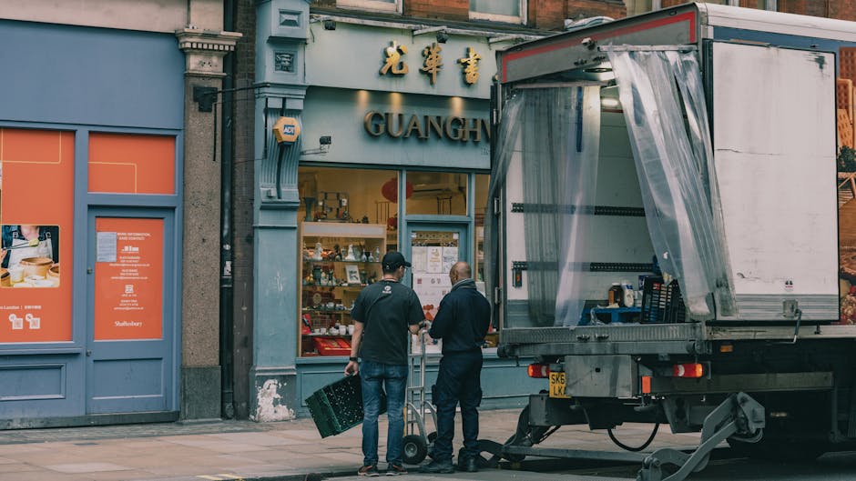 Two men are standing on a pavement in front of a shop with a grey and orange facade, engaged in a home relocation or moving process. One individual, wearing a black cap and dark clothing, holds a plastic crate, while the other, dressed more casually, is using a hand truck or dolly to transport a load. To their right, a partially open loading area of a box truck shows clear plastic wrapping covering some items inside. The scene is set outdoors during daytime, with the shop behind them displaying shop signage in Chinese characters and English, including the word 'GUANGH.' The loading process involves the careful handling of furniture or boxes, with the truck positioned close to the shop for easy access, reflecting typical procedures in furniture transport or packing and moving services, as offered by Man With a Van Wapping. The overall setting captures a typical moment in domestic removal logistics, focusing on loading or unloading activities outside a commercial property.