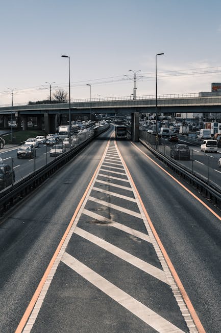 A wide view of a multi-lane urban road with a bridge overhead, showing vehicles including cars and vans in both directions. In the foreground, an area designated for loading or unloading is marked with diagonal white stripes and bordered by orange lines. On the pavement within this zone, several cardboard boxes, wrapped household belongings, and a few pieces of furniture are being carefully loaded or unloaded, with some items protected by blankets and plastic wrapping. A man with a van from Man With a Van Wapping is participating in a home relocation process, positioning items for transport. The scene takes place on a street with limited space, typical of narrow roads like Wapping Lane, illustrating the logistical challenges of moving in such environments. The lighting is natural, suggesting daytime, and the background shows a bridge structure and urban infrastructure, highlighting the need for precise planning in furniture transport and packing during a house move.