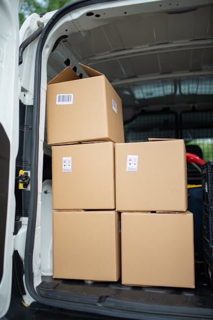Inside the rear cargo area of a white van operated by Man With a Van Wapping, four large cardboard boxes are neatly stacked, with two placed on the floor and two positioned on top, all secured with clear packing tape and white labels. The van's interior is illuminated by natural light from outside, revealing the vehicle's grey interior lining and a glimpse of other moving supplies, such as plastic wrap and blankets, stored nearby. The scene captures an organized loading process typical of home relocation or furniture transport services within the context of house removals, emphasizing careful packing and efficient use of space. The setting is outdoors, with the van's open rear door revealing the pavement and part of the surrounding environment. This image illustrates the logistical aspect of packing and loading boxes during a professional move, consistent with the services provided by [COMPANY_NAME] in coordinating efficient removal and transport operations for house relocations in the Wapping area, as referenced in the page about 'Tobacco Dock moves: Best van routes and loading spots.'