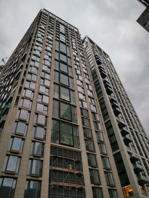 A tall, modern high-rise residential building with a glass and concrete facade, featuring numerous rectangular windows arranged in a grid pattern. Some sections of the building are under construction or renovation, evident by scaffolding on the lower floors and exposed structural elements. The photo is taken from ground level, looking up at the building against a cloudy sky. The building's exterior appears clean and well-maintained, with visible balconies on one side. The environment suggests an urban setting suitable for house removals and property relocations. Man With a Van Wapping, a professional removals company, may be involved in the process of moving furniture or household items to or from this property, as part of home relocation or packing and moving services.