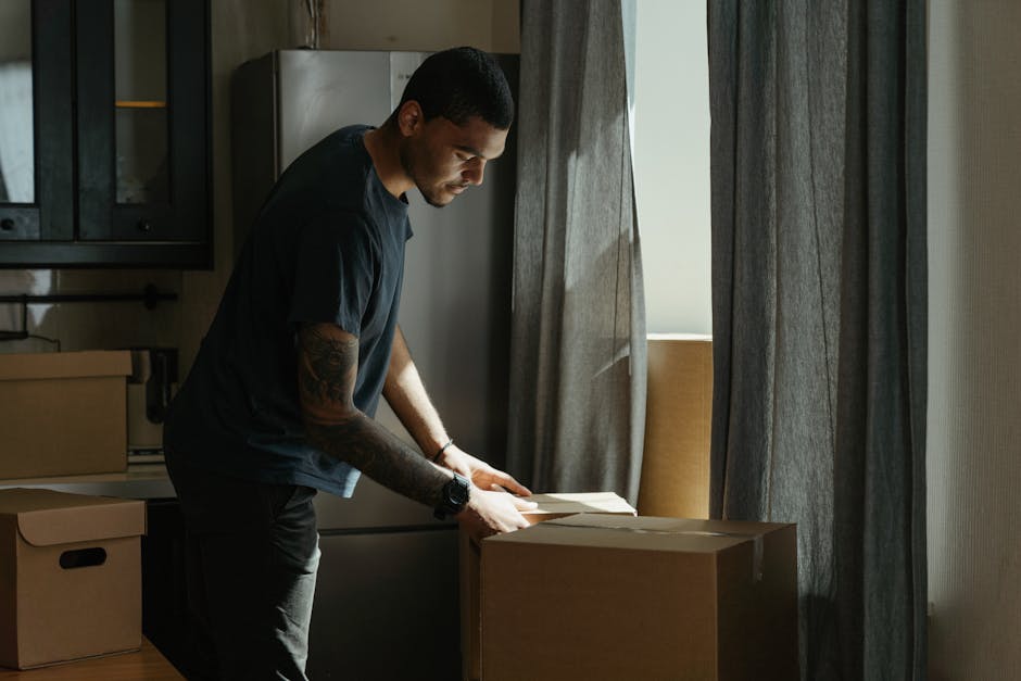 A man with dark hair and tattoos on his forearms is inside a residential kitchen, standing next to a stack of cardboard boxes. He is wearing a black t-shirt, black trousers, and a wristwatch, and is focused on packing or preparing items for a home relocation. The kitchen features dark cabinetry, a stainless steel refrigerator, and a window with sheer curtains allowing natural light to illuminate the scene. Some packing materials, such as bubble wrap or paper, may be visible on the boxes. The image captures the loading process as part of furniture transport related to a house moving or packing and moving service, with the man representing a professional from Man With a Van Wapping. The environment indicates an interior space actively being prepared for relocation, with organized packing taking place in readiness for transport, fitting the context of same-day access solutions for an urgent flat move in Wapping.