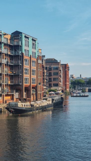 A row of modern, multi-story residential buildings with large glass windows and balconies, located along a riverbank on a clear day with blue skies. The buildings feature a mix of brick and metal cladding, some with visible outdoor furniture and potted plants on balconies. A black barge or houseboat is moored along the edge of the water close to the buildings. In the foreground, the calm river reflects the structures and sky, with gentle ripples on the surface. The scene depicts an urban waterfront environment suitable for home relocation and furniture transport activities, as provided by companies like Man With a Van Wapping, highlighting the logistical considerations of loading and unloading during removals near water-based locations.
